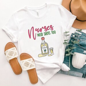 May include: White t-shirt with the text "Nurses need shots too" in pink and green lettering. A cartoon image of a bottle of tequila and a shot glass are below the text.