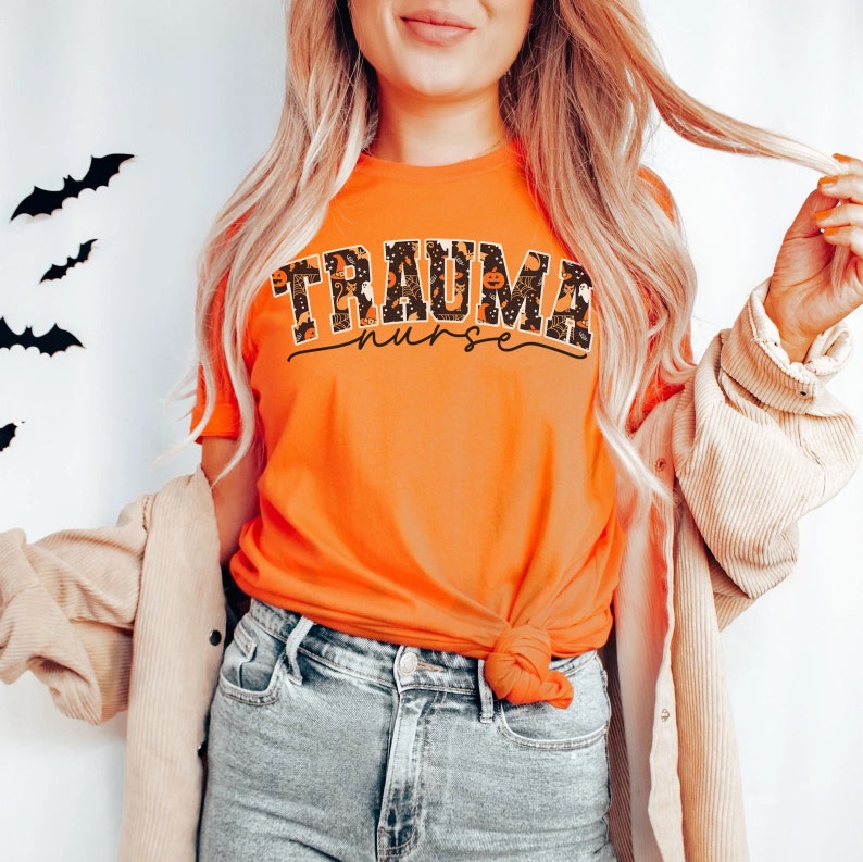 Puede incluir: Una camiseta naranja brillante con un estampado gr&aacute;fico negro y naranja que dice "Trauma Nurse" con im&aacute;genes de tem&aacute;tica de Halloween.