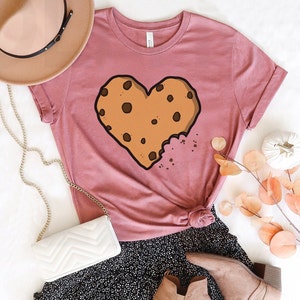 May include: A pink t-shirt with a brown chocolate chip cookie shaped like a heart. The cookie has a bite taken out of it.