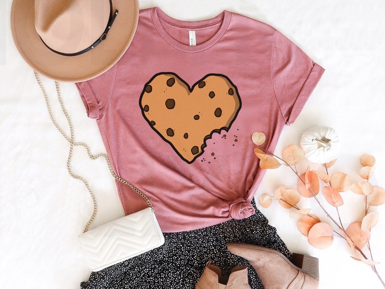 May include: A pink t-shirt with a brown chocolate chip cookie shaped like a heart. The cookie has a bite taken out of it.