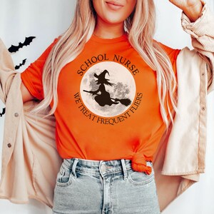 Puede incluir: Camiseta naranja con un gr&aacute;fico en blanco y negro de una bruja volando en una escoba frente a una luna llena. El gr&aacute;fico est&aacute; rodeado de un c&iacute;rculo con el texto "School Nurse We Treat Frequent Flyers".