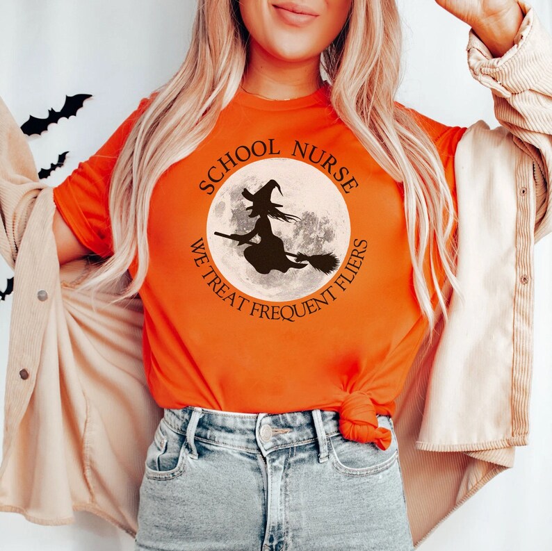 Puede incluir: Camiseta naranja con un gr&aacute;fico en blanco y negro de una bruja volando en una escoba frente a una luna llena. El gr&aacute;fico est&aacute; rodeado de un c&iacute;rculo con el texto "School Nurse We Treat Frequent Flyers".