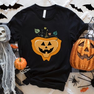 May include: Black t-shirt with a graphic of a smiling jack-o'-lantern with a stem and leaves. The shirt is surrounded by Halloween decorations, including a spider web, bats, and candy.