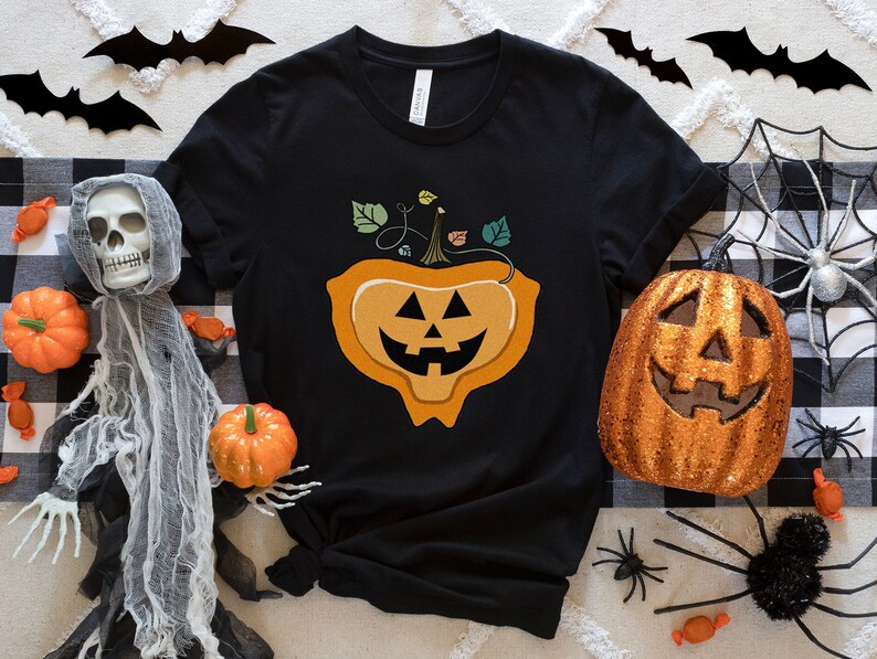 May include: Black t-shirt with a graphic of a smiling jack-o'-lantern with a stem and leaves. The shirt is surrounded by Halloween decorations, including a spider web, bats, and candy.