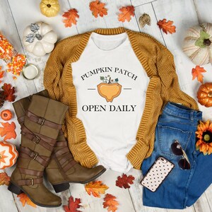 K&ouml;nnte beinhalten: Ein wei&szlig;es T-Shirt mit dem Text "Pumpkin Patch - Open Daily" und einer Grafik mit einem K&uuml;rbis in einem Korb. Das Shirt liegt auf einer wei&szlig;en Holzoberfl&auml;che mit einem braunen Cardigan, einer blauen Jeans, braunen Stiefeln und Herbstdekorationen.