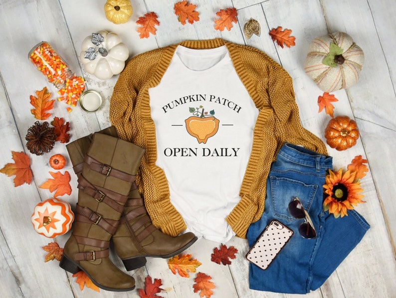 K&ouml;nnte beinhalten: Ein wei&szlig;es T-Shirt mit dem Text "Pumpkin Patch - Open Daily" und einer Grafik mit einem K&uuml;rbis in einem Korb. Das Shirt liegt auf einer wei&szlig;en Holzoberfl&auml;che mit einem braunen Cardigan, einer blauen Jeans, braunen Stiefeln und Herbstdekorationen.