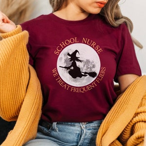 Puede incluir: Una camiseta color granate con un gr&aacute;fico circular blanco que presenta una bruja volando en una escoba frente a una luna llena. El texto alrededor del c&iacute;rculo dice "School Nurse We Treat Frequent Flyers".