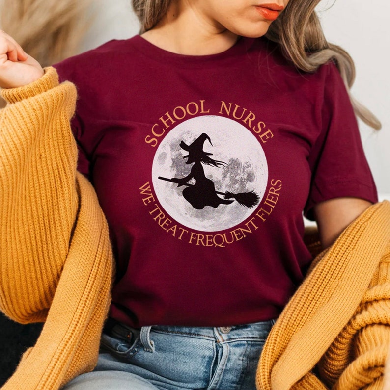 Puede incluir: Una camiseta color granate con un gr&aacute;fico circular blanco que presenta una bruja volando en una escoba frente a una luna llena. El texto alrededor del c&iacute;rculo dice "School Nurse We Treat Frequent Flyers".