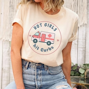 May include: A beige t-shirt featuring a graphic of a red ambulance with a white cross and the text "HOT GIRLS hit CURBS" in a circle with pink hearts.