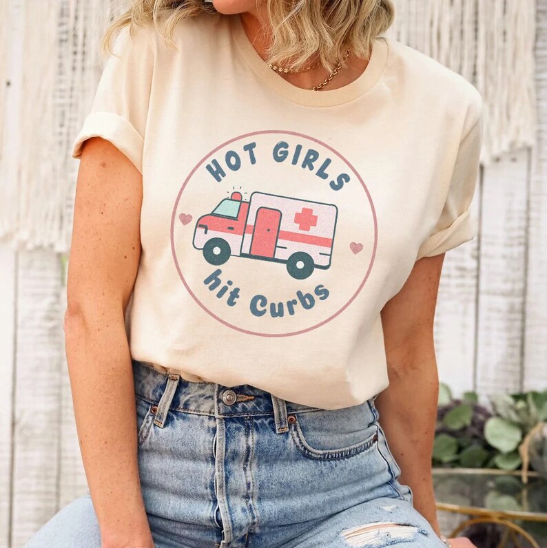 May include: A beige t-shirt featuring a graphic of a red ambulance with a white cross and the text "HOT GIRLS hit CURBS" in a circle with pink hearts.