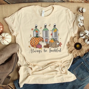 May include: A beige t-shirt with a graphic design of three scuba tanks, pumpkins, and fall leaves. The text "Always be Tankful" is printed below the graphic.