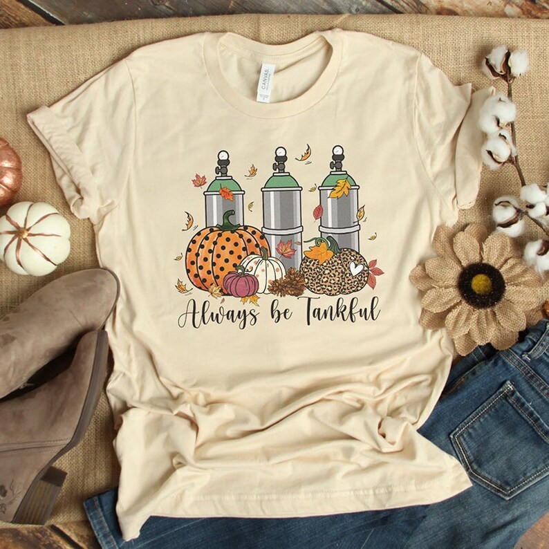 May include: A beige t-shirt with a graphic design of three scuba tanks, pumpkins, and fall leaves. The text "Always be Tankful" is printed below the graphic.