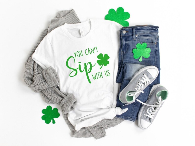 May include: White t-shirt with green shamrock and the text "You Can't Sip With Us". The shirt is laid out on a white surface with a gray sweater, blue jeans, and green shamrock decorations.