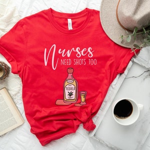 May include: Red t-shirt with the text "Nurses need shots too" and a cartoon illustration of a bottle of tequila and a shot glass.