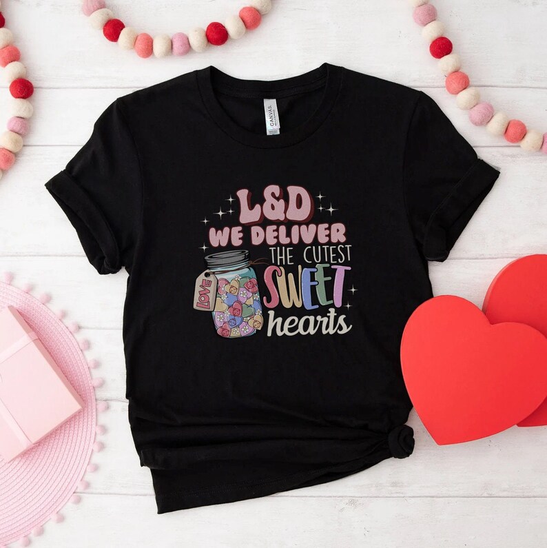 May include: Black t-shirt with a graphic of a jar filled with colorful hearts and the text "L&D We Deliver The Cutest Sweet Hearts".
