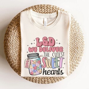 May include: A beige t-shirt with the text "L&D We Deliver The Cutest Sweet Hearts" in pink, blue, and yellow. A mason jar filled with colorful candy hearts is pictured next to the text.