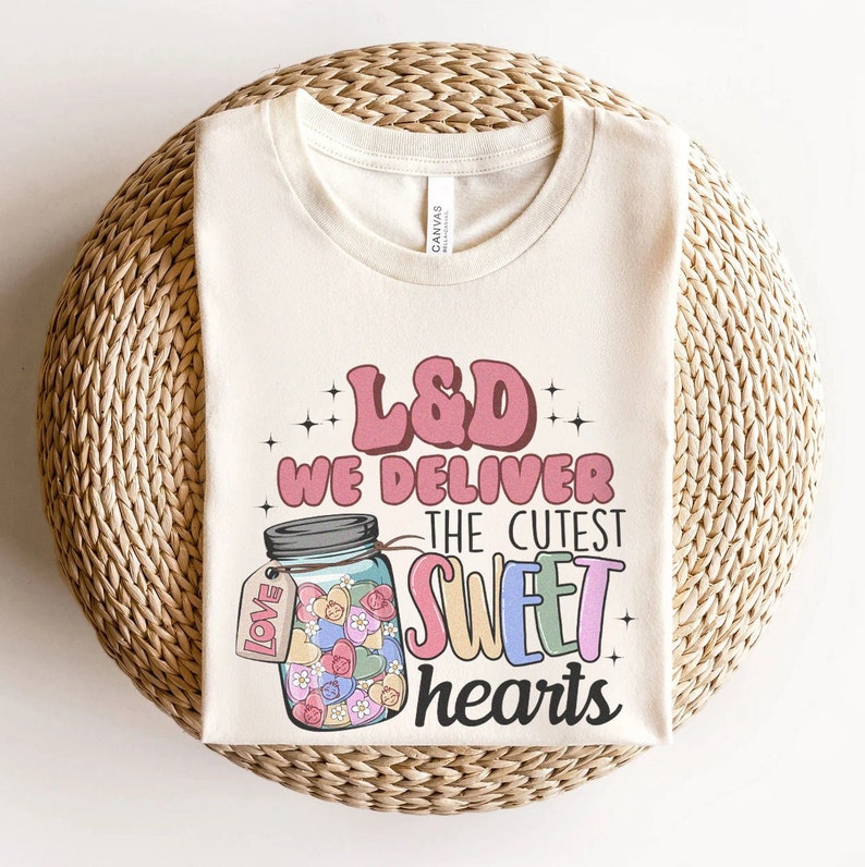 May include: A beige t-shirt with the text "L&D We Deliver The Cutest Sweet Hearts" in pink, blue, and yellow. A mason jar filled with colorful candy hearts is pictured next to the text.