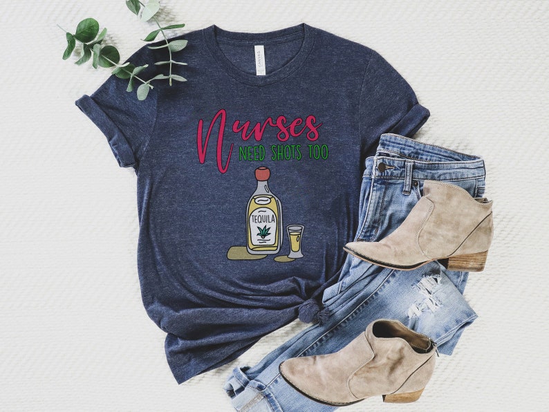 May include: A navy blue t-shirt with the text "Nurses need shots too" in red and green. A cartoon image of a bottle of tequila and a shot glass is below the text.
