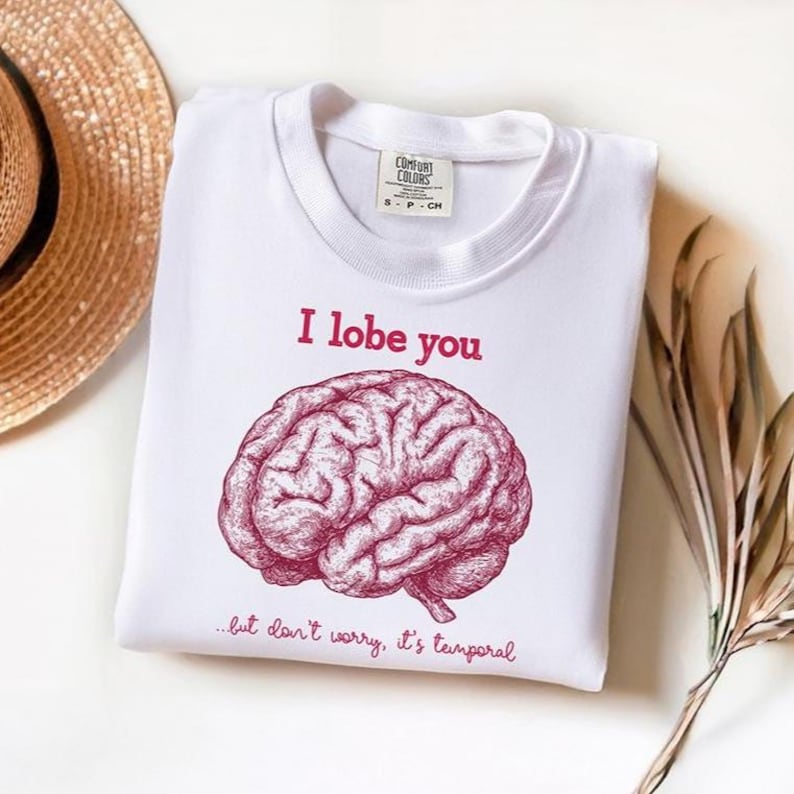 May include: White crew neck sweatshirt featuring the burgundy text "I lobe you" and a brain illustration. The phrase "...but don't worry, it's temporal" is below the brain graphic. A Comfort Colors label is visible.
