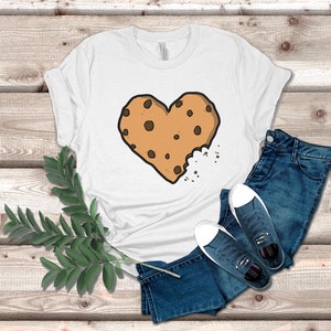 May include: White t-shirt with a brown chocolate chip cookie in the shape of a heart. The cookie is slightly bitten into, with crumbs scattered around it.