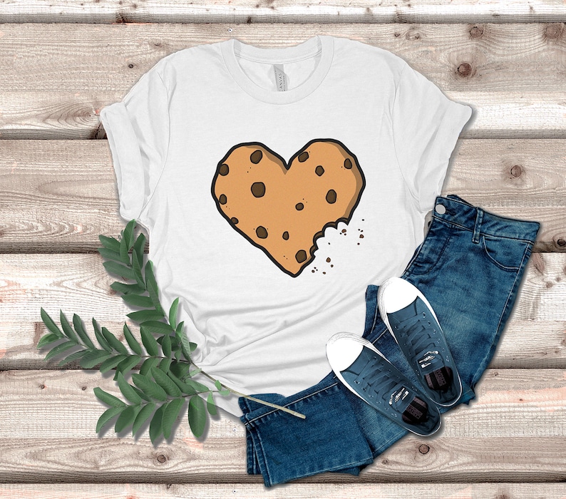 May include: White t-shirt with a brown chocolate chip cookie in the shape of a heart. The cookie is slightly bitten into, with crumbs scattered around it.