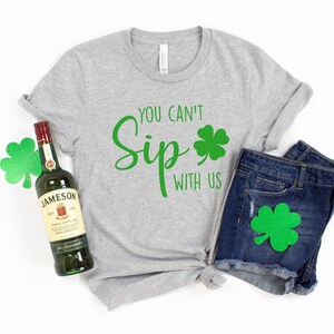 May include: A gray t-shirt with the text "You can't Sip with us" and a green shamrock. A bottle of Jameson Irish Whiskey and a pair of blue denim shorts with a green shamrock on the pocket.