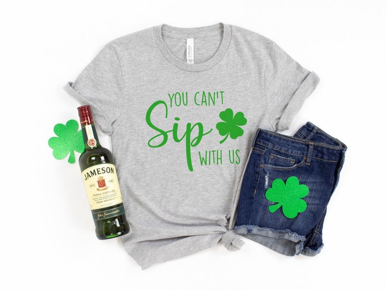 May include: A gray t-shirt with the text "You can't Sip with us" and a green shamrock. A bottle of Jameson Irish Whiskey and a pair of blue denim shorts with a green shamrock on the pocket.