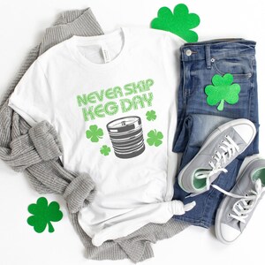 May include: White t-shirt with the text "Never Skip Keg Day" in green letters and a green illustration of a keg. The shirt is laid out on a white surface with a gray sweater, blue jeans, and green shamrock decorations.
