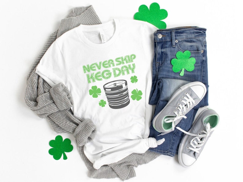 May include: White t-shirt with the text "Never Skip Keg Day" in green letters and a green illustration of a keg. The shirt is laid out on a white surface with a gray sweater, blue jeans, and green shamrock decorations.