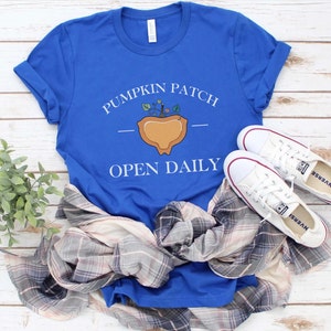 K&ouml;nnte beinhalten: Ein k&ouml;nigsblaues T-Shirt mit einer wei&szlig;en Grafik eines K&uuml;rbisfeldes mit dem Text "Pumpkin Patch" und "Open Daily". Das T-Shirt liegt auf einer wei&szlig;en Holzoberfl&auml;che mit einem karierten Hemd und wei&szlig;en Turnschuhen.