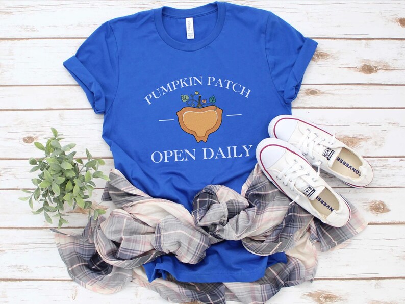 K&ouml;nnte beinhalten: Ein k&ouml;nigsblaues T-Shirt mit einer wei&szlig;en Grafik eines K&uuml;rbisfeldes mit dem Text "Pumpkin Patch" und "Open Daily". Das T-Shirt liegt auf einer wei&szlig;en Holzoberfl&auml;che mit einem karierten Hemd und wei&szlig;en Turnschuhen.