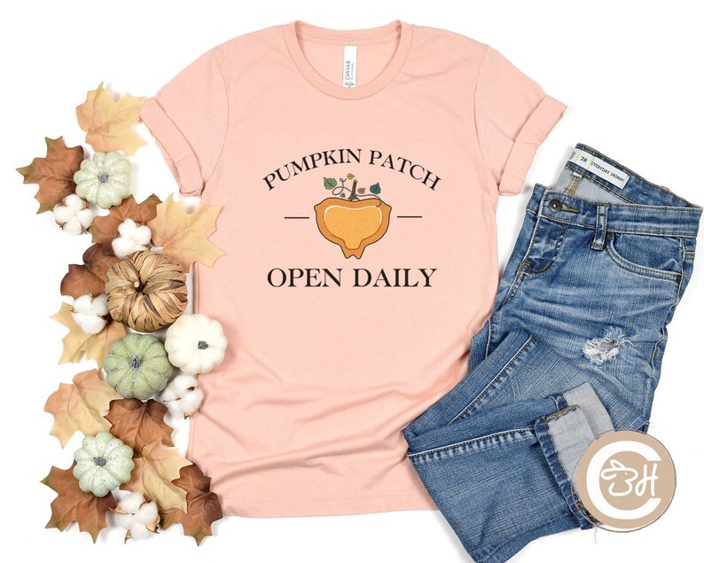 K&ouml;nnte beinhalten: Ein pinkfarbenes T-Shirt mit dem Text "Pumpkin Patch - Open Daily" und einer Grafik einer K&uuml;rbis mit Bl&auml;ttern. Das Shirt liegt auf einer wei&szlig;en Oberfl&auml;che mit Herbstdekorationen.