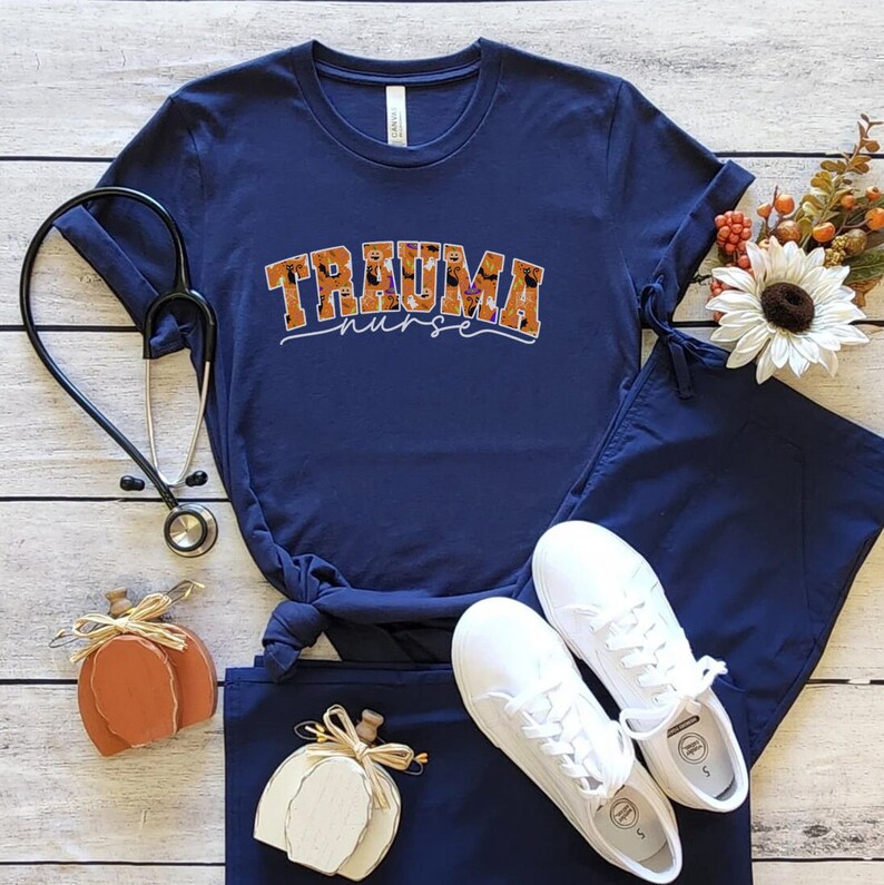 Puede incluir: Una camiseta azul marino con el texto "Trauma Nurse" en letras naranja y negras. Las letras est&aacute;n decoradas con un patr&oacute;n de calabazas naranja y negras. La camiseta se combina con pantalones de trabajo azul marino y zapatillas blancas.