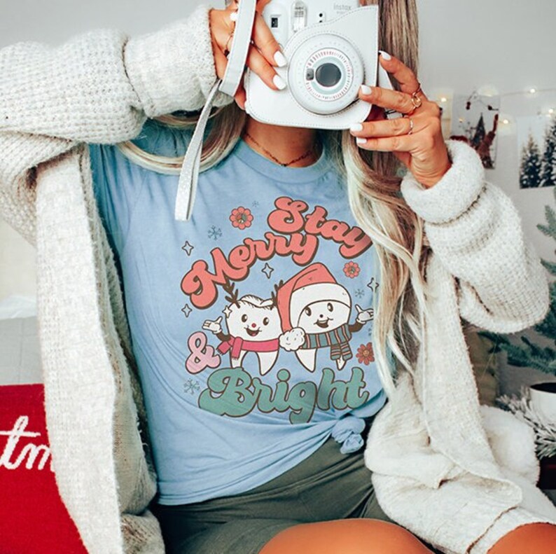 May include: A light blue t-shirt with a Christmas themed graphic featuring two cartoon teeth. The teeth are wearing Santa hats and scarves. The text on the shirt reads "Stay Merry & Bright".