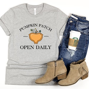 K&ouml;nnte beinhalten: Ein graues T-Shirt mit dem Text "Pumpkin Patch Open Daily" und einer stilisierten K&uuml;rbisgrafik.