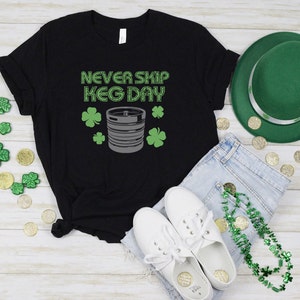 May include: Black t-shirt with the text "Never Skip Keg Day" in white letters. A silver keg is pictured below the text with green shamrocks on either side.