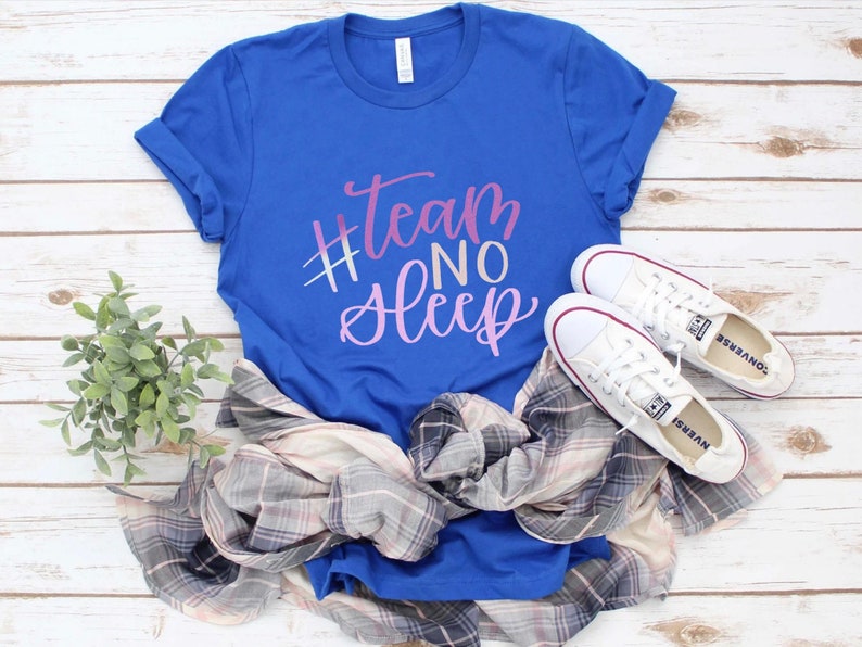 May include: A royal blue t-shirt with the text "#Team No Sleep" in a gradient pink and purple font. The shirt is paired with a plaid shirt, and white Converse sneakers. The items are arranged on a white wooden surface.