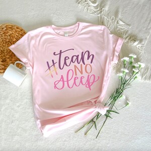 May include: A light pink t-shirt with the text "#Team No Sleep" in a gradient of purple, orange, and pink. The shirt is folded and tied at the bottom. A white mug and flowers are in the background.