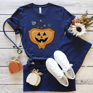 May include: A navy blue scrub top with a pumpkin jack-o'-lantern design. The pumpkin has a smiling face with a green stem and leaves. The scrub top is paired with matching navy blue scrub pants and white sneakers.