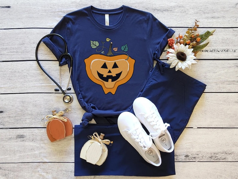 May include: A navy blue scrub top with a pumpkin jack-o'-lantern design. The pumpkin has a smiling face with a green stem and leaves. The scrub top is paired with matching navy blue scrub pants and white sneakers.