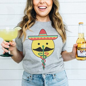 K&ouml;nnte beinhalten: Ein graues T-Shirt mit einem gelben Smiley, der einen bunten Sombrero und einen gr&uuml;n-roten medizinischen Caduceus tr&auml;gt. Die Person h&auml;lt eine Margarita und eine Flasche Corona Light Bier.