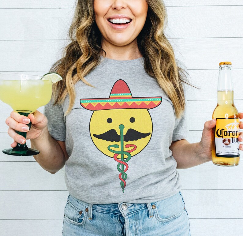 K&ouml;nnte beinhalten: Ein graues T-Shirt mit einem gelben Smiley, der einen bunten Sombrero und einen gr&uuml;n-roten medizinischen Caduceus tr&auml;gt. Die Person h&auml;lt eine Margarita und eine Flasche Corona Light Bier.