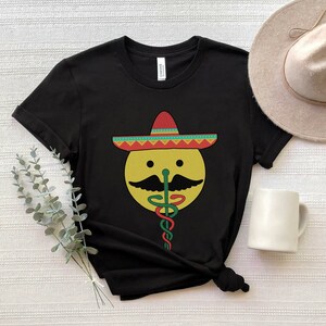 K&ouml;nnte beinhalten: Schwarzes T-Shirt mit einem gelben Smiley, der einen roten, gelben und gr&uuml;nen Sombrero tr&auml;gt. Der Smiley hat einen Schnurrbart und eine gr&uuml;ne Schlange als Nase.