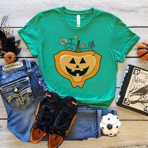May include: A green t-shirt with a graphic of a melting jack-o'-lantern. The shirt is laying on a wooden surface with jeans, sneakers, a book, and a small pumpkin.