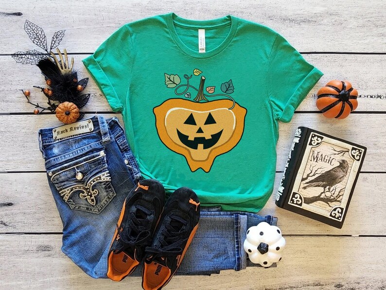 May include: A green t-shirt with a graphic of a melting jack-o'-lantern. The shirt is laying on a wooden surface with jeans, sneakers, a book, and a small pumpkin.