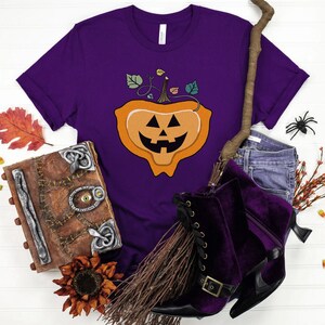 May include: Purple t-shirt with a jack-o'-lantern graphic. The shirt is surrounded by Halloween-themed props, including a broom, a book, candy corn, and fall leaves.