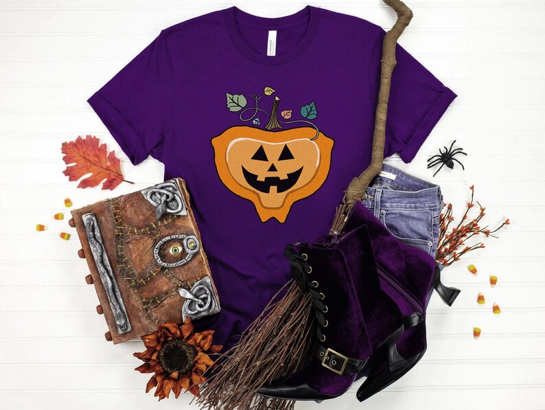 May include: Purple t-shirt with a jack-o'-lantern graphic. The shirt is surrounded by Halloween-themed props, including a broom, a book, candy corn, and fall leaves.