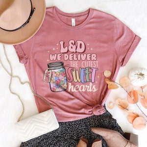 May include: A pink t-shirt with the text "L&D We Deliver The Cutest Sweet Hearts" and a graphic of a mason jar filled with colorful hearts. The shirt is styled with a white purse, a brown hat, and a black and white skirt.