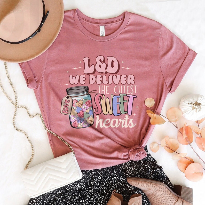 May include: A pink t-shirt with the text "L&D We Deliver The Cutest Sweet Hearts" and a graphic of a mason jar filled with colorful hearts. The shirt is styled with a white purse, a brown hat, and a black and white skirt.