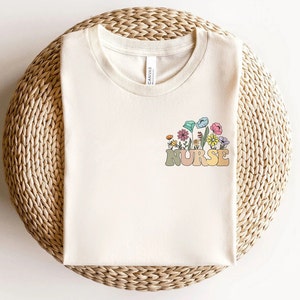 May include: Cream-colored t-shirt with a floral design and the word "NURSE" in a retro font. The flowers are in shades of pink, blue, yellow, and orange. The shirt is made of a soft material and is perfect for everyday wear.